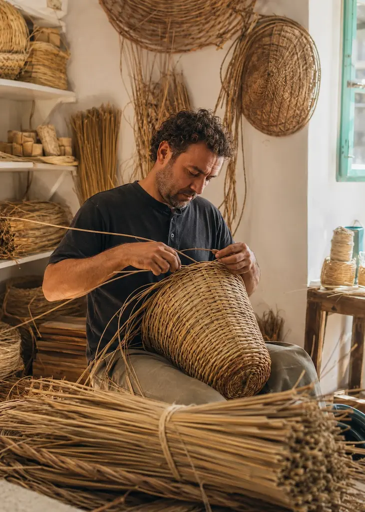 Artisan weaving a large basket by hand from natural plant fibers inside a traditional sunlit workshop.