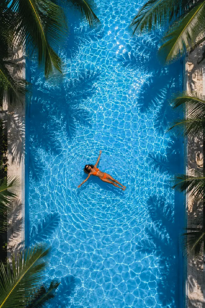 Aerial view of a woman floating in a blue swimming pool surrounded by palm trees