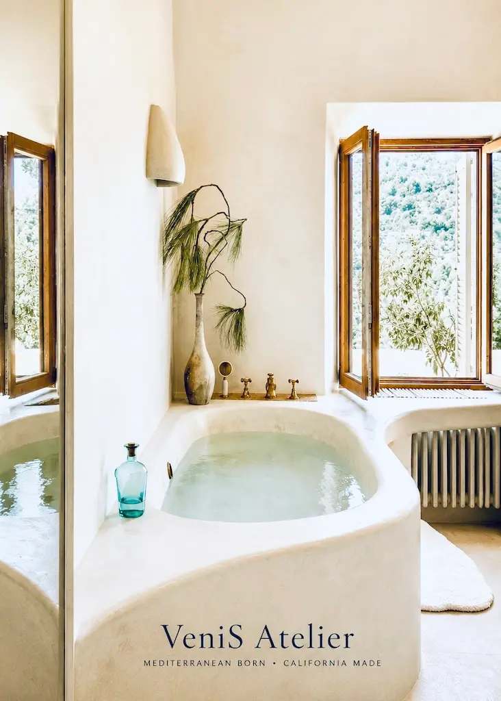 Rounded plaster bathtub beside open wooden windows, with brass taps, a ceramic vase and leafy branches in natural light.