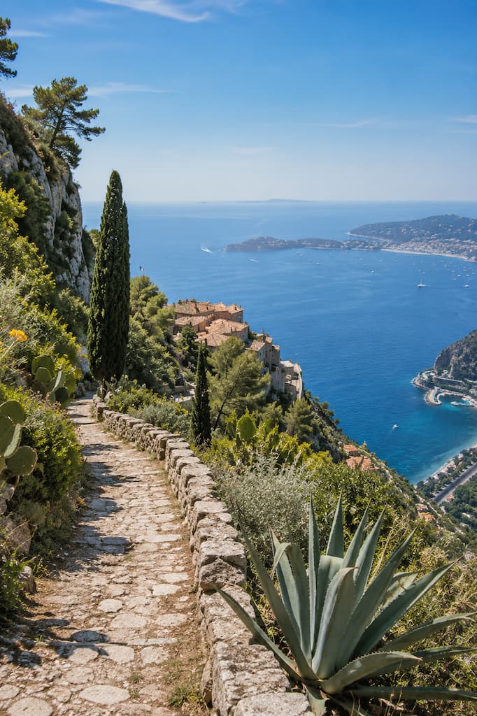 Stone path bordered by agaves and cypress trees above a hillside village and blue sea.