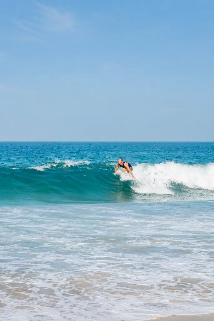 A surfer rides a turquoise ocean wave near shore beneath a clear blue sky.