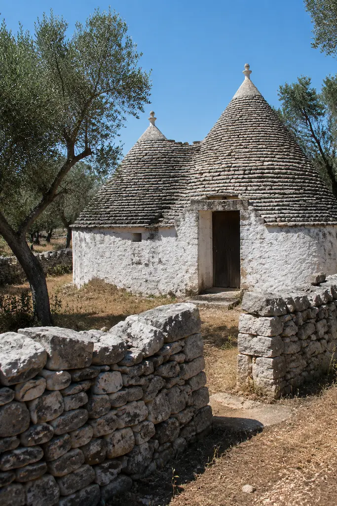 Whitewashed trullo with conical stone roofs, framed by olive trees and a dry-stone wall.