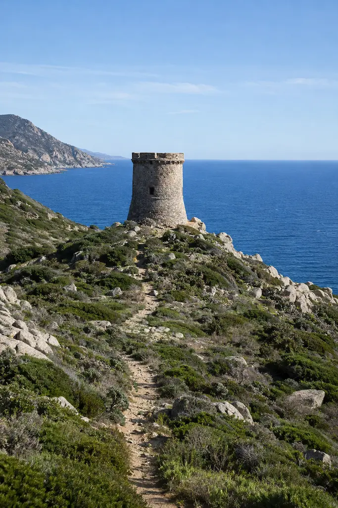 Stone coastal watchtower on a rocky headland above the Mediterranean Sea, with a narrow path through low shrubs.