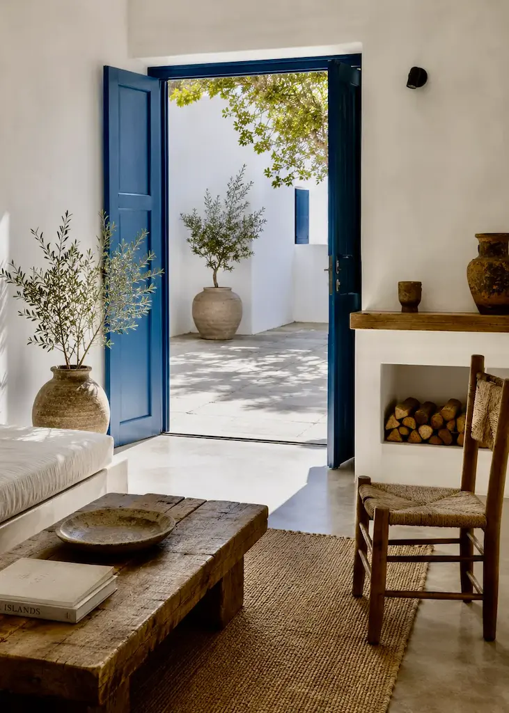 Simple Greek home with built-in plaster benches, colorful linen cushions, bougainvillea, natural wood table, and a lounging cat illustrating contemporary Greek interior design.