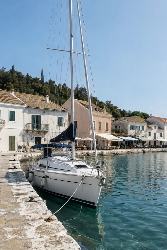 White sailboat moored beside a stone quay, with pastel waterfront houses and clear blue-green harbour water.