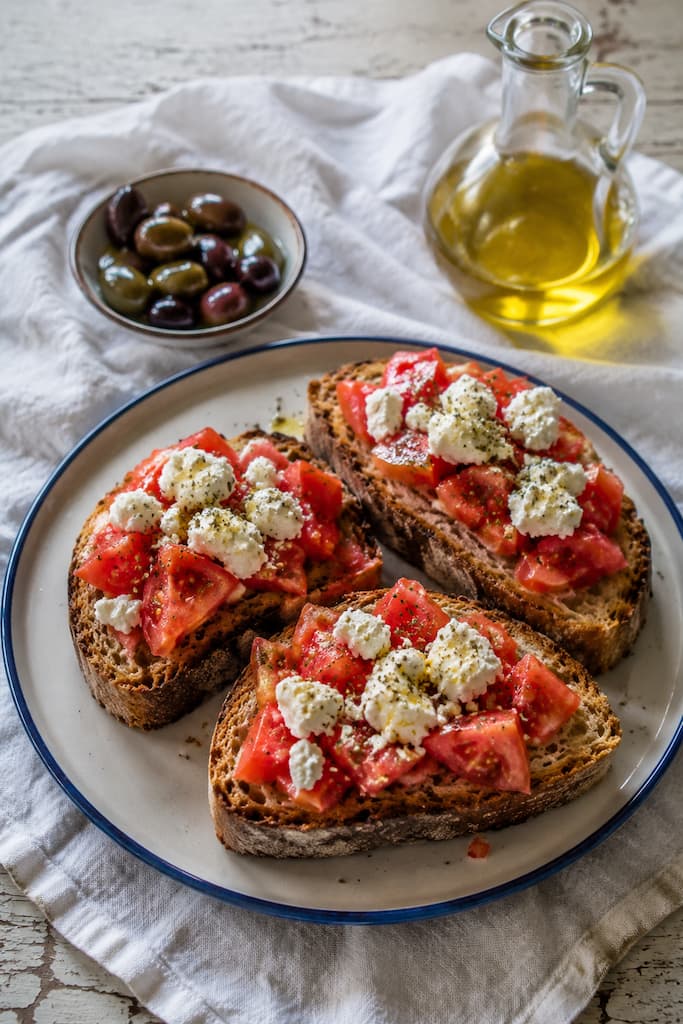 Rustic bread topped with chopped tomatoes, crumbled white cheese, and oregano, beside a glass jug of olive oil and a bowl of olives.