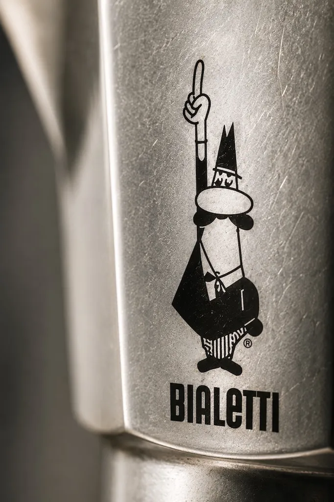 Close-up of the iconic Bialetti mustached man logo on a classic aluminum Moka Express coffee maker.