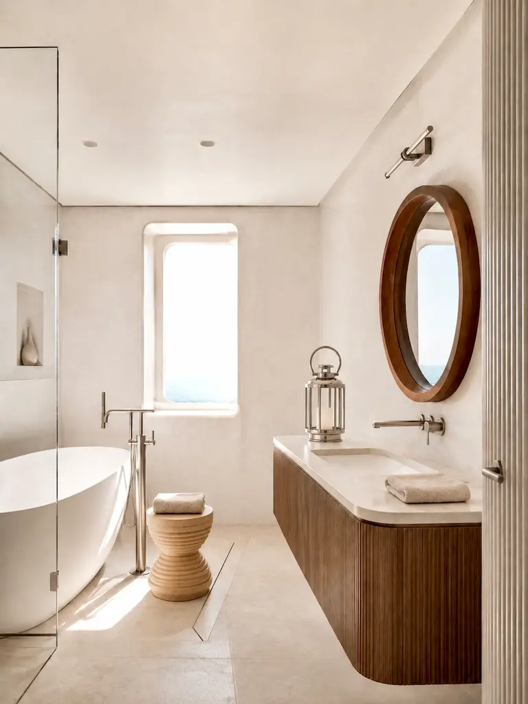 Minimal yacht-inspired bathroom with curved wood vanity, round mirror, freestanding tub and ocean view through a porthole-style window.