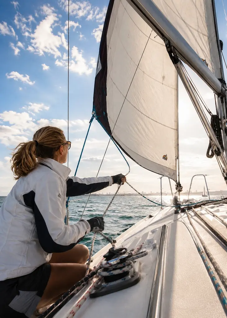 A sailor beginner adjusts a line while trimming the sail aboard a sailboat on open water.