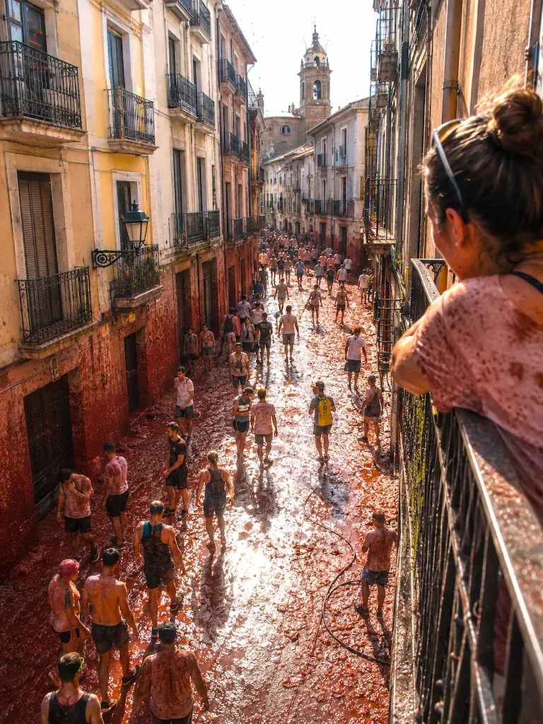 A woman leans over a balcony as festivalgoers walk through a tomato-covered street in Buñol after La Tomatina, with a historic bell tower in the distance.