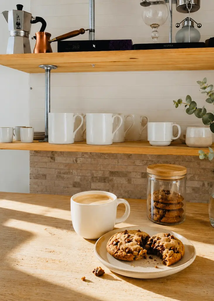 Craft coffee and warm chocolate chip cookies on a bright California kitchen counter with natural wood shelves and morning sunlight.