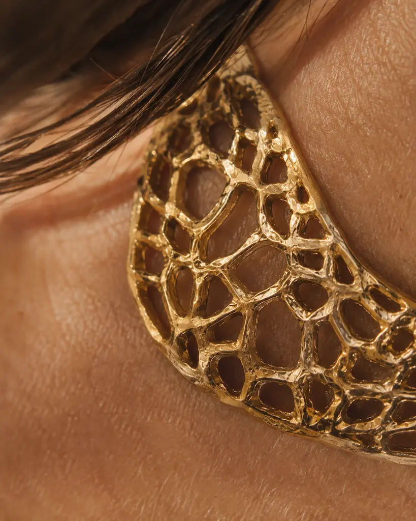 Extreme close-up of handmade openwork gold jewelry against skin, showing irregular pierced cells, fine scratches and texture.