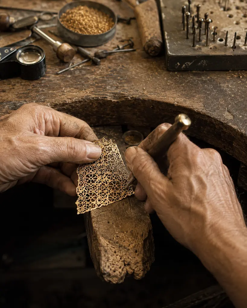 Jeweler working a delicate pierced gold sheet by hand at a worn wooden bench surrounded by traditional tools and metal filings.