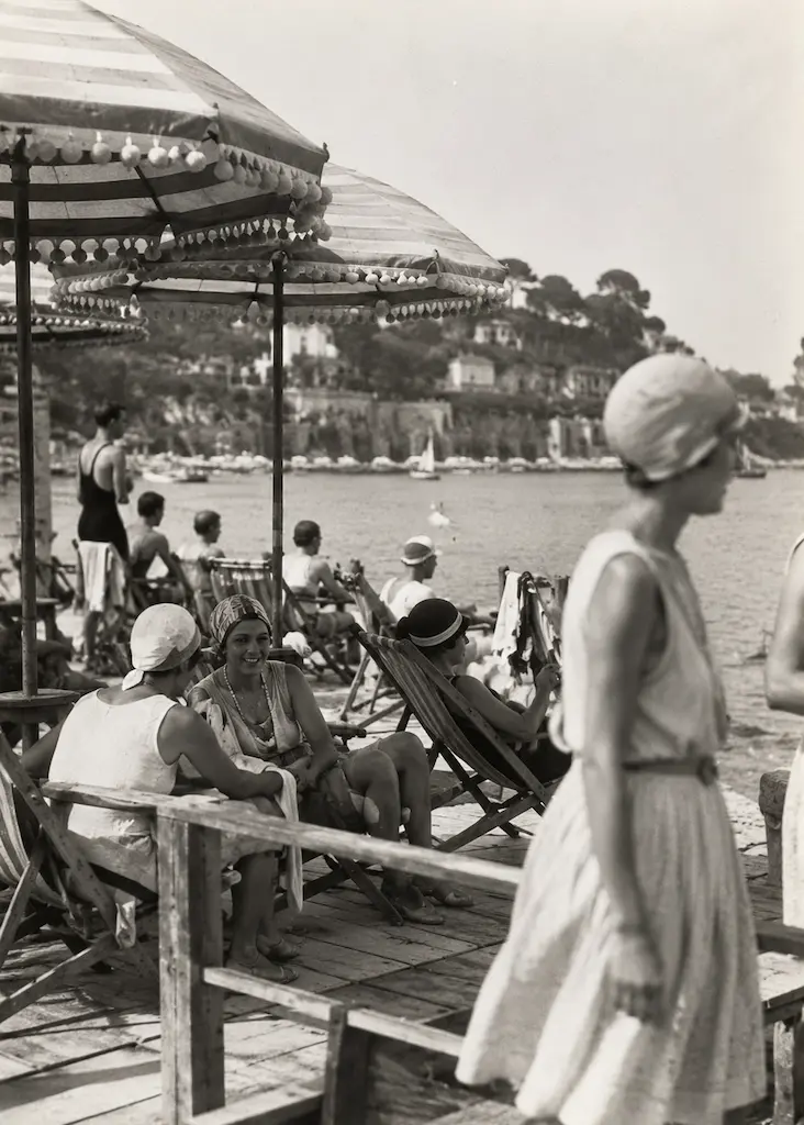 Beachgoers relaxing beneath striped parasols at a French Riviera seaside club in the early 20th century.