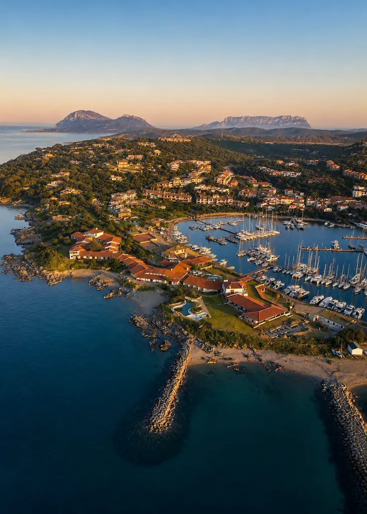Aerial view of Porto Rotondo, Sardinia, with its marina, terracotta-roofed village, rocky coastline and surrounding Mediterranean landscape.