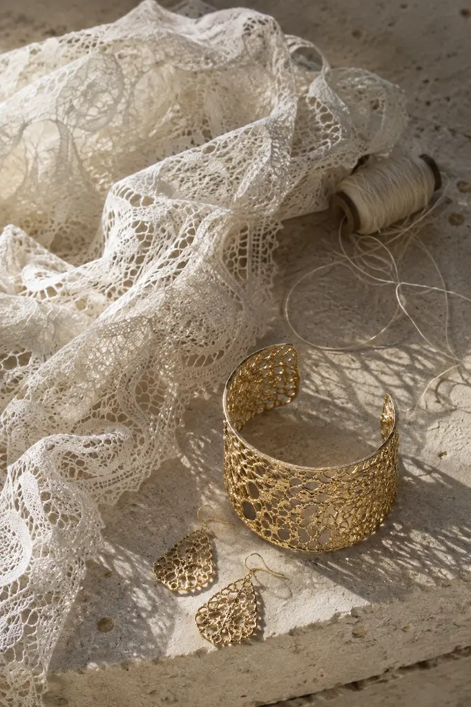 Fine white lace, loose thread and an openwork gold cuff arranged on sunlit stone, their shadows blending textile and metal patterns.