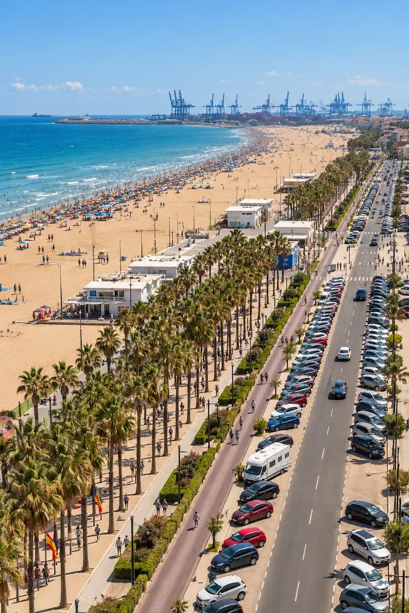 Elevated view of Malvarrosa Beach in Valencia, Spain, showing golden sand, turquoise Mediterranean waters, a palm-lined promenade, and the Port of Valencia in the distance.
