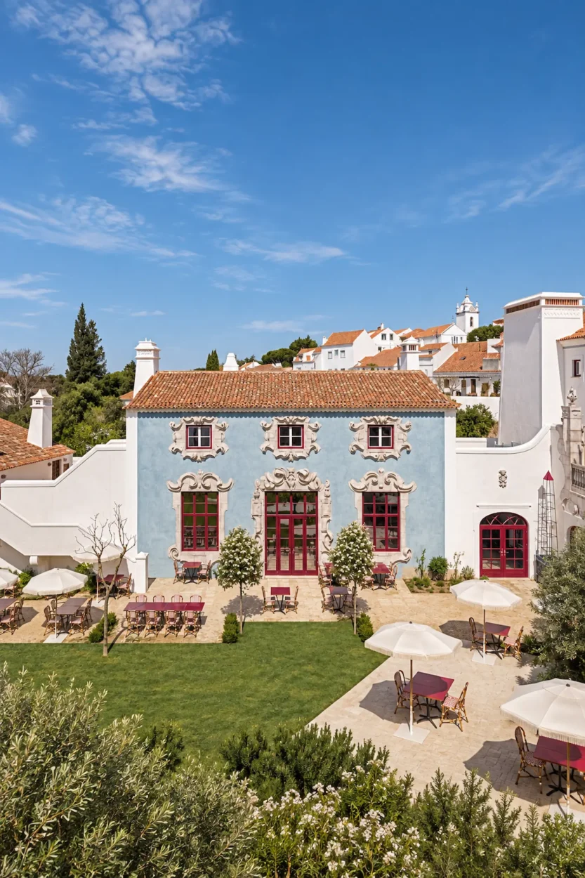 Exterior view of Vermelho in Melides, Portugal—a pale blue boutique hotel with ornate stone window surrounds and red-framed doors, facing a sunlit courtyard with café tables, white parasols, manicured lawn and Mediterranean greenery beneath a clear blue sky.